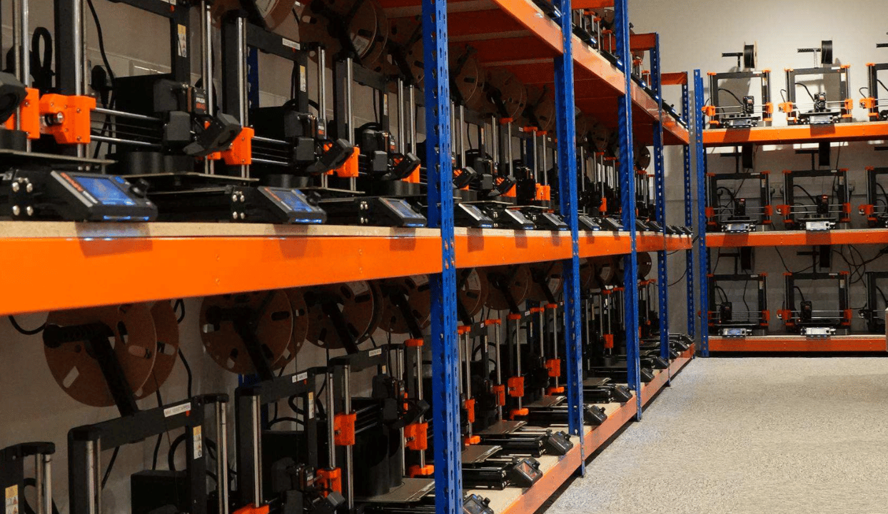 A 3D print farm with dozens of desktop FDM printers lined up in rows on orange-and-blue industrial shelving, each fed by a cardboard filament spool mounted above it, in a warehouse-style room.