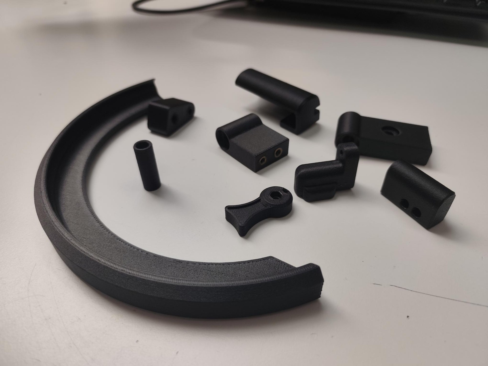 Eight black end-use parts 3D printed in rPA12 + 10% recycled carbon fiber, laid out on a white desk: a large curved arc segment in the foreground and a set of smaller brackets, clamps, a spacer tube and a lever arm with bolt holes.
